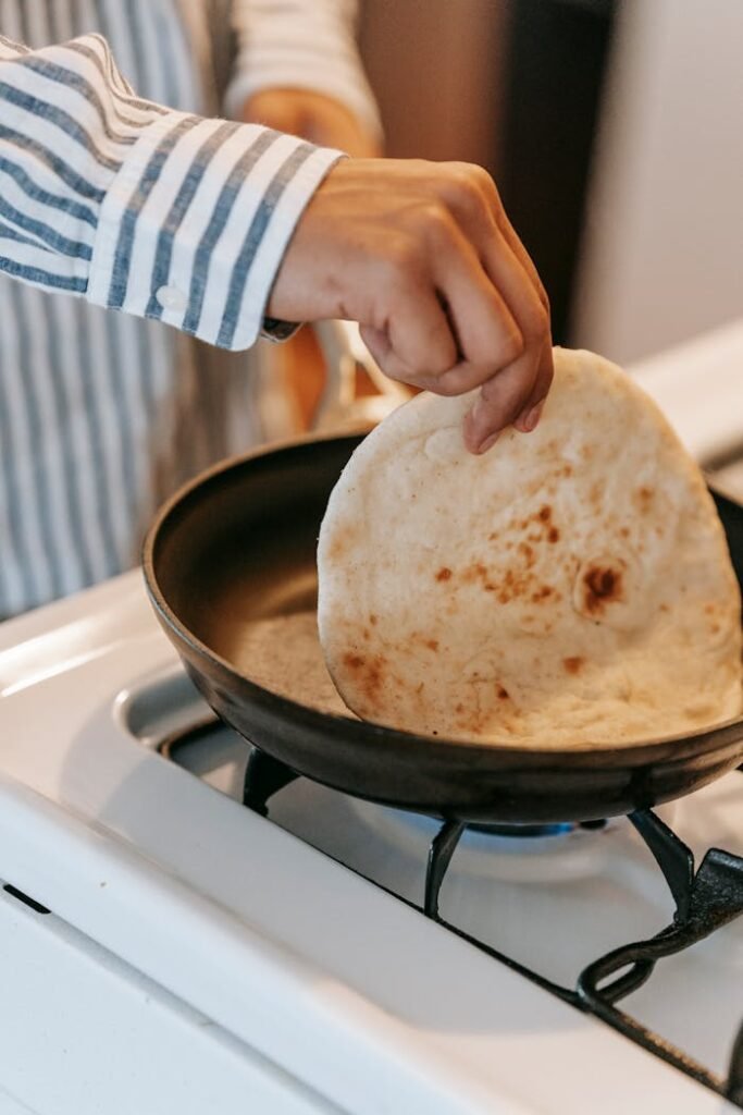 Crop anonymous male cook preparing traditional Indian chapati bread in pan placed on stove in kitchen at home